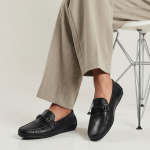 Mocassins en cuir noir avec bride tressée et boucle métallique, portés pieds nus avec un pantalon fluide gris clair. La posture est assise, jambes croisées, sur une chaise design blanche aux pieds métalliques croisés, dans un studio au fond neutre et lumineux.