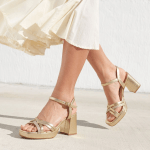 Sandales à talons hauts blocs et plateformes dorés métallisés avec de fines brides croisées à l'avant, portées avec une jupe ou robe fluide blanc cassé. La posture est en mouvement de marche, capturée de profil sous une lumière estivale sur un sol en béton clair devant un mur texturé blanc.